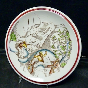 May include: A white ceramic plate with a red rim features a vintage illustration of three figures in a mountainous landscape. The figures are dressed in brown and yellow clothing and are carrying rifles. The scene is depicted in a brown and green color palette.