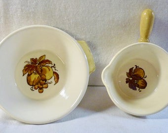 May include: Two white ceramic bowls with a fruit design. The larger bowl has a yellow rim and a handle. The smaller bowl has a yellow handle.