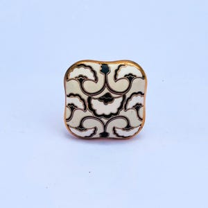 May include: Square decorative cabinet knob with a floral design. The knob features a cream-colored background with black and white floral patterns. It has a gold-colored trim around the edges.