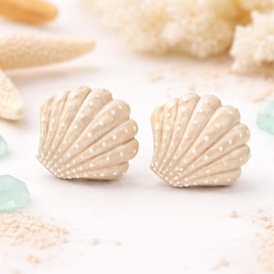 May include: Two beige seashell-shaped decorative objects with white dotted patterns. The shells are on a white surface with sand and other beach-themed elements in the background. The shells are likely used for home decor.