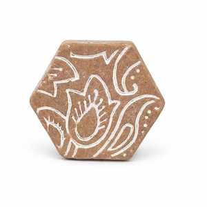 Op de afbeelding: Zeshoekige, bruine decoratieve knop met een wit bloemmotief. De knop heeft een textuur en is waarschijnlijk gemaakt van steen of een vergelijkbaar materiaal. Het witte ontwerp is een gedetailleerd, gestileerd bloemenpatroon.