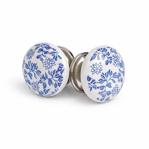 Könnte beinhalten: Zwei weiße Keramik-Türknöpfe mit blauem Blumenmuster. Die Knöpfe haben eine runde Form und sind auf silberfarbenen Metallbasen montiert. Das florale Design zeigt stilisierte Blumen und Blätter.