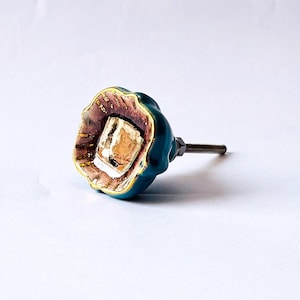 May include: A decorative cabinet knob with a teal exterior and a gold-trimmed interior. The knob has a square, gold-colored center and a threaded rod for installation. The knob's interior is a mix of brown and gold.
