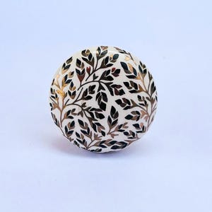 Może przedstawiać: Okrągła gałka do szafki w kolorze złamanej bieli z botanicznym wzorem. Wzór przedstawia ciemnobrązowe i złote liście i gałęzie. Gałka jest elementem dekoracyjnym mebli.