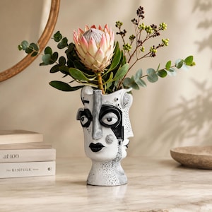 May include: A white ceramic vase shaped like a human face with black painted details, including eyes, lips, and abstract patterns. The vase holds a large pink protea flower, green eucalyptus branches, and small brown berries.