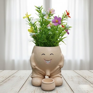 May include: A whimsical beige ceramic planter shaped like a person, with a smiling face and arms. The planter holds a vibrant arrangement of artificial flowers in various colors. A small matching pot sits in front.