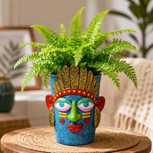 May include: A blue planter shaped like a face with a green nose, red lips, and a gold feather headdress, containing a vibrant green fern plant. The face features large blue eyes with pink eyelids and red circular cheek accents on a textured blue surface.