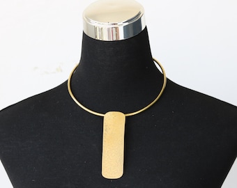 Peut inclure: Un collier doré avec un pendentif rectangulaire. Le collier est composé d'un collier circulaire rigide et d'un pendentif allongé texturé. Le bijou est présenté sur un mannequin noir sur fond blanc.