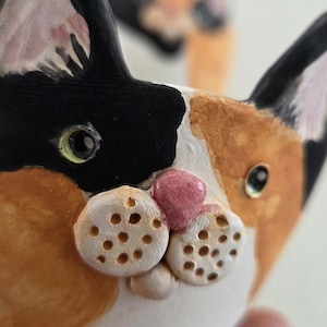 May include: A ceramic bowl shaped like a calico cat head with black, orange, and white patches. The cat features a pink nose, textured white muzzle with small indentations for whiskers, and two pointed ears with black and pink painted details.