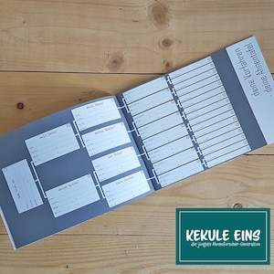 Könnte beinhalten: Ein offenes Stammbaum-Buch mit grauem Einband und weißen Innenseiten. Die Tabelle enthält Felder zum Ausfüllen von Familiengeschichtsdetails. Der Text "KEKULE EINS" und "die jüngste Ahnenforscher-Generation" befindet sich in einem grünen Feld.