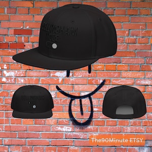 May include: Black flat-brimmed baseball cap with the text "BELLINGHAM MODE:" in raised lettering. The cap has a white button on the front and a flat brim. The cap is displayed against a brick wall.