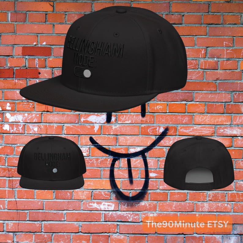 May include: Black flat-brimmed baseball cap with the text "BELLINGHAM MODE:" in raised lettering. The cap has a white button on the front and a flat brim. The cap is displayed against a brick wall.