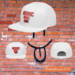 May include: White baseball cap with a flat brim. The cap features the text "MODRIC MODE:" in red and blue. The cap is displayed against a brick wall background.