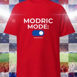 K&ouml;nnte beinhalten: Ein rotes T-Shirt mit dem wei&szlig;en Schriftzug "MODRIC MODE:" &uuml;ber einer blau-wei&szlig;en Toggle-Grafik. Der Text "THE90MINUTE" befindet sich darunter. Das T-Shirt wird vor dem Hintergrund eines Fu&szlig;ballstadions pr&auml;sentiert.