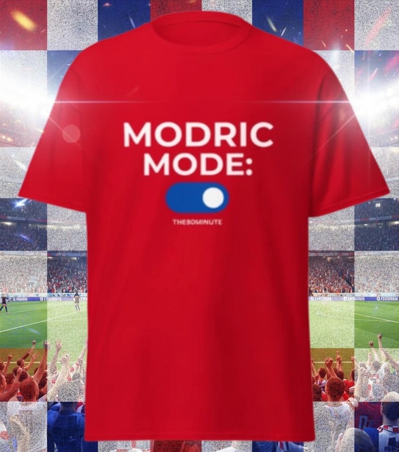K&ouml;nnte beinhalten: Ein rotes T-Shirt mit dem wei&szlig;en Schriftzug "MODRIC MODE:" &uuml;ber einer blau-wei&szlig;en Toggle-Grafik. Der Text "THE90MINUTE" befindet sich darunter. Das T-Shirt wird vor dem Hintergrund eines Fu&szlig;ballstadions pr&auml;sentiert.