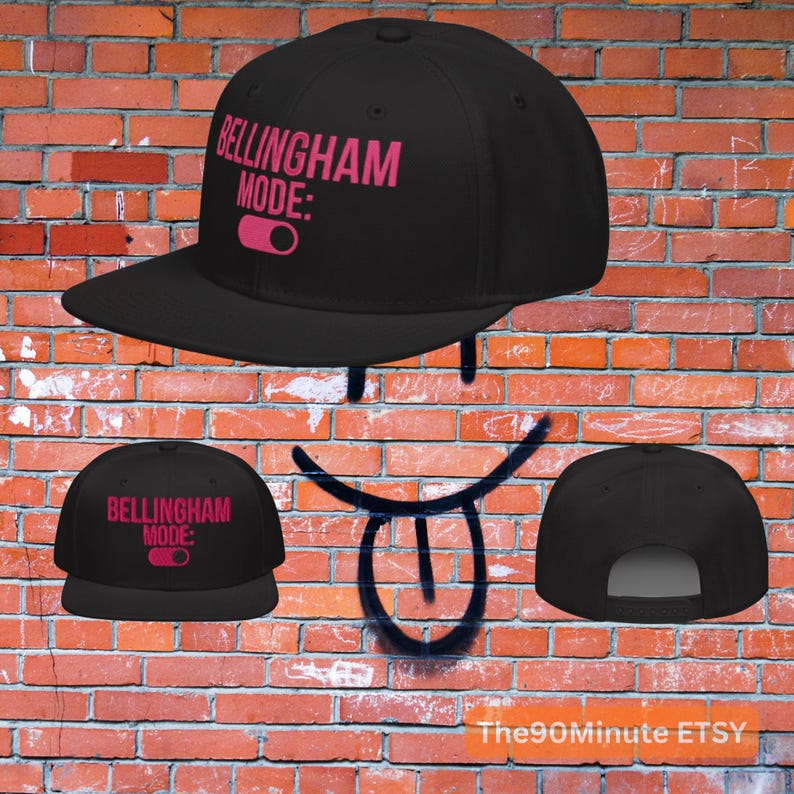 May include: Black baseball cap with the text "Bellingham Mode:" in pink. The cap has a flat brim and a snapback closure. The design includes a pink toggle graphic.