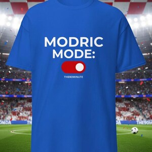 K&ouml;nnte beinhalten: K&ouml;nigsblaues T-Shirt mit dem wei&szlig;en Text "MODRIC MODE:" &uuml;ber einer rot-wei&szlig;en Toggle-Grafik. Der Text "THE90MINUTE" befindet sich darunter. Der Hintergrund zeigt ein Fu&szlig;ballfeld.