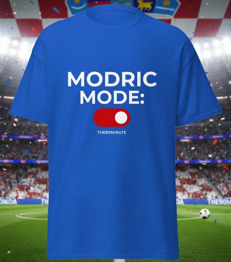 K&ouml;nnte beinhalten: K&ouml;nigsblaues T-Shirt mit dem wei&szlig;en Text "MODRIC MODE:" &uuml;ber einer rot-wei&szlig;en Toggle-Grafik. Der Text "THE90MINUTE" befindet sich darunter. Der Hintergrund zeigt ein Fu&szlig;ballfeld.
