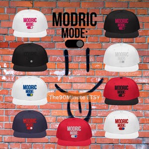 May include: A collection of snapback baseball caps in various colors, including white, black, red, and navy blue. Each cap features the text "MODRIC MODE" with a toggle graphic. The caps are displayed against a brick wall background.