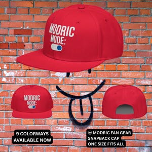 May include: A red snapback cap with the text "MODRIC MODE:" in white. The cap has a flat brim and a gray underside. The image also shows the text "9 COLORWAYS AVAILABLE NOW" and "MODRIC FAN GEAR SNAPBACK CAP ONE SIZE FITS ALL".