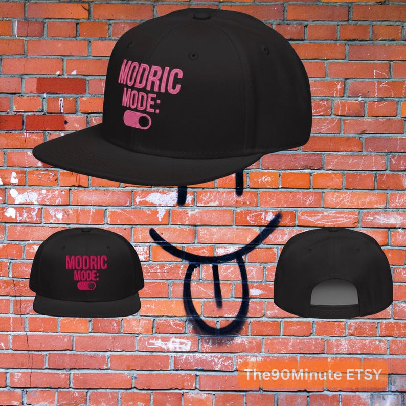 May include: Black flat-brimmed baseball cap with the text "MODRIC MODE:" in pink. The cap features a pink toggle graphic. The cap is displayed against a brick wall background.