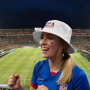 May include: A white bucket hat with the letters "USA" printed on the front in red and blue. The hat is worn by a person at a football match, with the American flag painted on their cheek. The person is wearing a blue shirt with the USA logo.