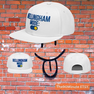 May include: White baseball cap with the text "BELLINGHAM MODE:" in blue. The design includes a blue and yellow toggle switch graphic. The cap has a flat brim and a snapback closure.