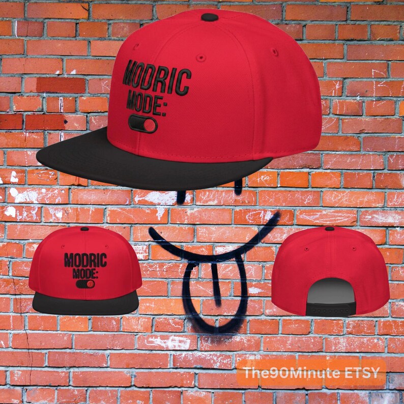 May include: A red baseball cap with a black brim. The cap features the text "MODRIC MODE:" in black, along with a graphic of a switch. The cap is displayed against a brick wall background.