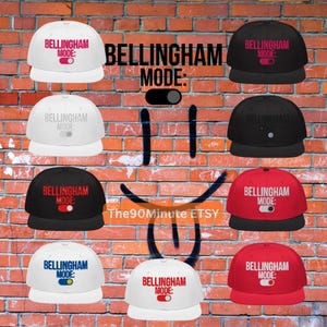 May include: A collection of baseball caps in various colors, including white, black, and red. Each cap features the text "BELLINGHAM MODE:" with a toggle graphic. The caps are displayed against a brick wall background.