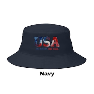 May include: A navy blue bucket hat featuring the word "USA" in red, white, and blue, with the text "ONE NATION. ONE TEAM." The hat is a casual style, suitable for sun protection. The word "Navy" is printed at the bottom of the image.