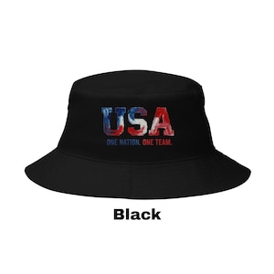 May include: Black bucket hat featuring the word "USA" in red, white, and blue, with the text "ONE NATION. ONE TEAM." The hat has a wide brim and a classic design.