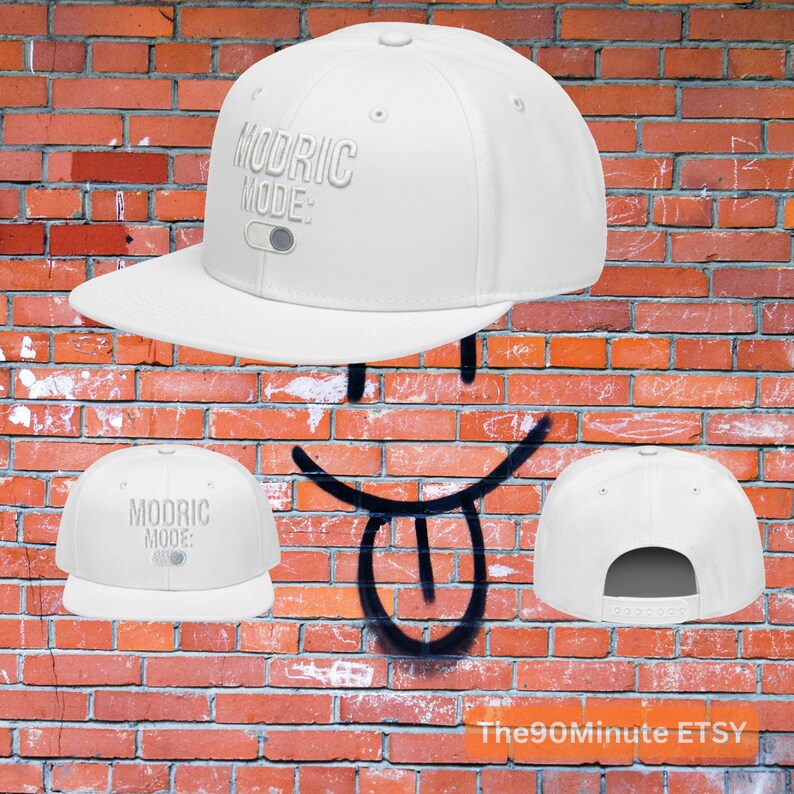 May include: A white flat-brimmed baseball cap with the text "MODRIC MODE:" in a light gray color. The cap has a snapback closure and a curved brim. The background is a red brick wall.