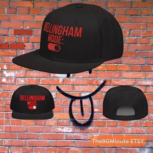 May include: Black baseball cap with the text "Bellingham Mode:" in red. The cap features a flat brim and a snapback closure. The design includes a red toggle graphic. The background is a brick wall.