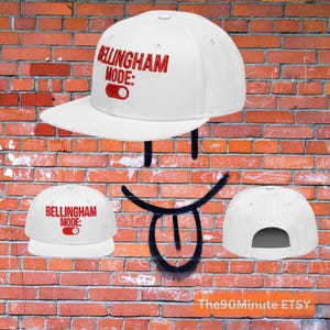 May include: White baseball cap with the text "BELLINGHAM MODE:" in red. The cap has a flat brim and an adjustable snapback closure. The design includes a red toggle graphic.