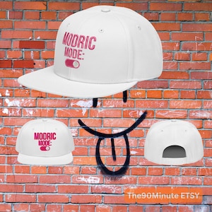 May include: White baseball cap with a flat brim. The cap features the text "MODRIC MODE:" in pink, along with a pink power button graphic. The cap is set against a brick wall background.