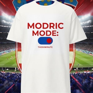 K&ouml;nnte beinhalten: Wei&szlig;es T-Shirt mit dem Text "MODRIC MODE:" in Rot, einer blau-roten Grafik und dem Text "THE90MINUTE" in Wei&szlig;. Das Shirt ist vor einem Stadionhintergrund abgebildet.