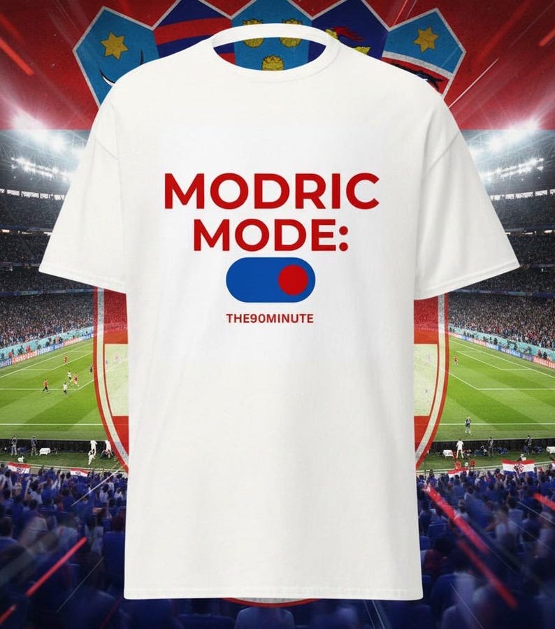 K&ouml;nnte beinhalten: Wei&szlig;es T-Shirt mit dem Text "MODRIC MODE:" in Rot, einer blau-roten Grafik und dem Text "THE90MINUTE" in Wei&szlig;. Das Shirt ist vor einem Stadionhintergrund abgebildet.