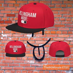 May include: A red and black snapback hat with the text "BELLINGHAM MODE:" in white. The hat has a black brim and a black button on top. The hat is displayed against a brick wall background.