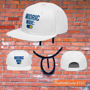 May include: White baseball cap with the text "MODRIC MODE:" in blue, along with a blue and yellow graphic. The cap has a flat brim and a snapback closure. The background is a brick wall.