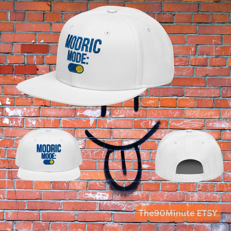 May include: White baseball cap with the text "MODRIC MODE:" in blue, along with a blue and yellow graphic. The cap has a flat brim and a snapback closure. The background is a brick wall.