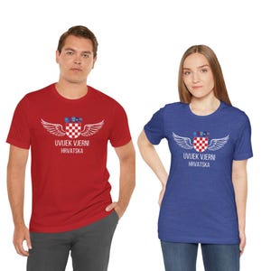 May include: A red short-sleeved t-shirt and a blue short-sleeved t-shirt, both featuring a white graphic of a checkered shield with wings and the text UVIJEK VJERNI HRVATSKA printed in white on the chest area of each garment.