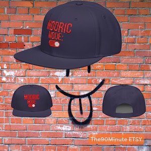 May include: Navy blue baseball cap with the text "MODRIC MODE:" in red. The cap features a flat brim and an adjustable strap. The design includes a red toggle switch graphic.