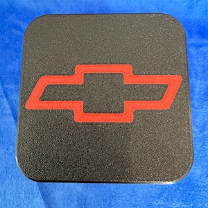 May include: A black square coaster with rounded corners and a red Chevrolet logo. The coaster has a textured surface. The background is blue.
