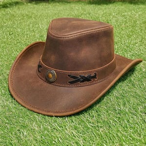May include: A brown leather cowboy hat with a decorative band and a circular emblem. The hat has a classic brim and a structured crown. The band features black stitched accents. The hat is displayed on a green grassy surface.