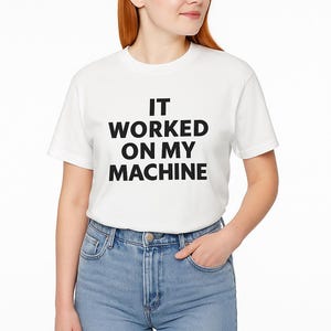 Peut inclure: T-shirt blanc avec le texte "IT WORKED ON MY MACHINE" en lettres noires grasses. Le t-shirt est porté avec un jean bleu. Le modèle a les cheveux roux.
