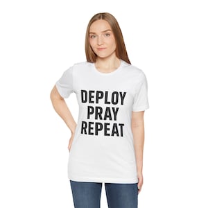 Könnte beinhalten: Weißes T-Shirt mit dem Text "DEPLOY PRAY REPEAT" in fetten schwarzen Buchstaben. Das Shirt hat kurze Ärmel und einen Rundhalsausschnitt. Die Person trägt blaue Jeans.