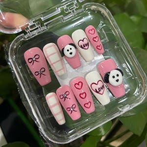 May include: A set of pink and white press-on nails with various designs. Some nails feature bows, hearts, and the word "Love." Others have a panda design or a checkered pattern. The nails are in a clear plastic case.