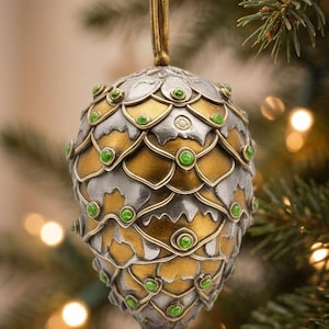 May include: Ornate Christmas ornament with a gold ribbon. The ornament is shaped like a pinecone, with silver and gold metal scales and green jewel accents. The background is a blurred Christmas tree with lights.