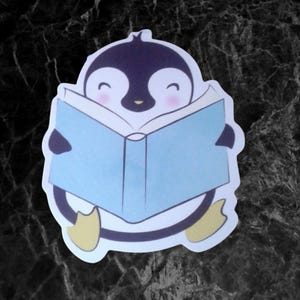 May include: A sticker featuring a cartoon penguin reading a light blue book. The penguin has a black body, white belly, and yellow feet. Its face is white with pink cheeks and a small yellow beak. The penguin is smiling.