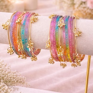 Könnte beinhalten: Ein Set bunter Armreifen mit einem Regenbogenverlauf aus Rosa, Orange, Gelb, Grün und Blau. Jeder Armreif ist mit goldfarbenen Akzenten und kleinen, hängenden Charms verziert. Der Schmuck wird auf einem weißen Ständer präsentiert.